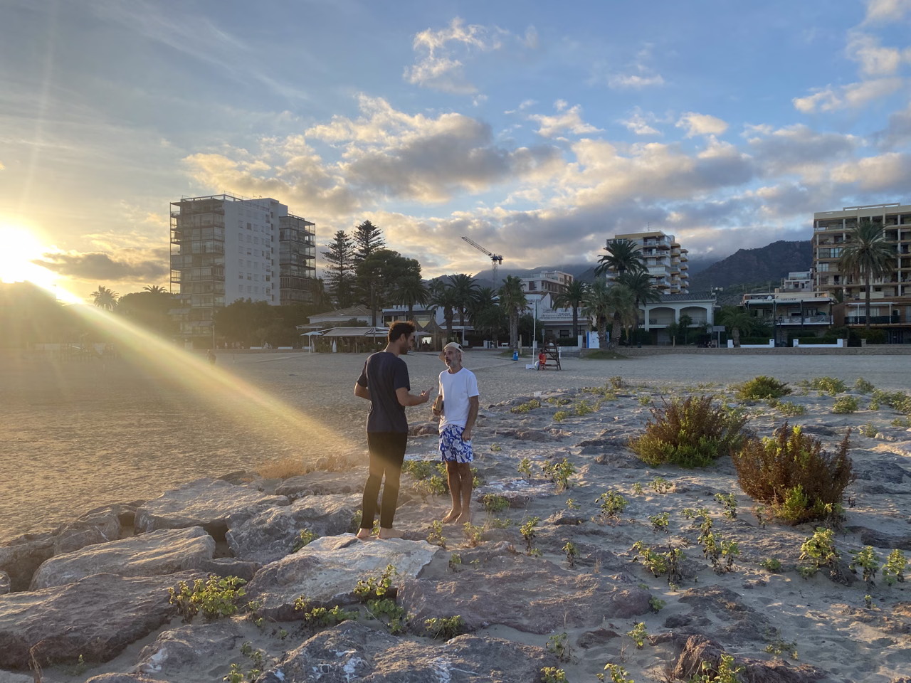 Two people walk across a beach toward apartment buildings in the evening sun.